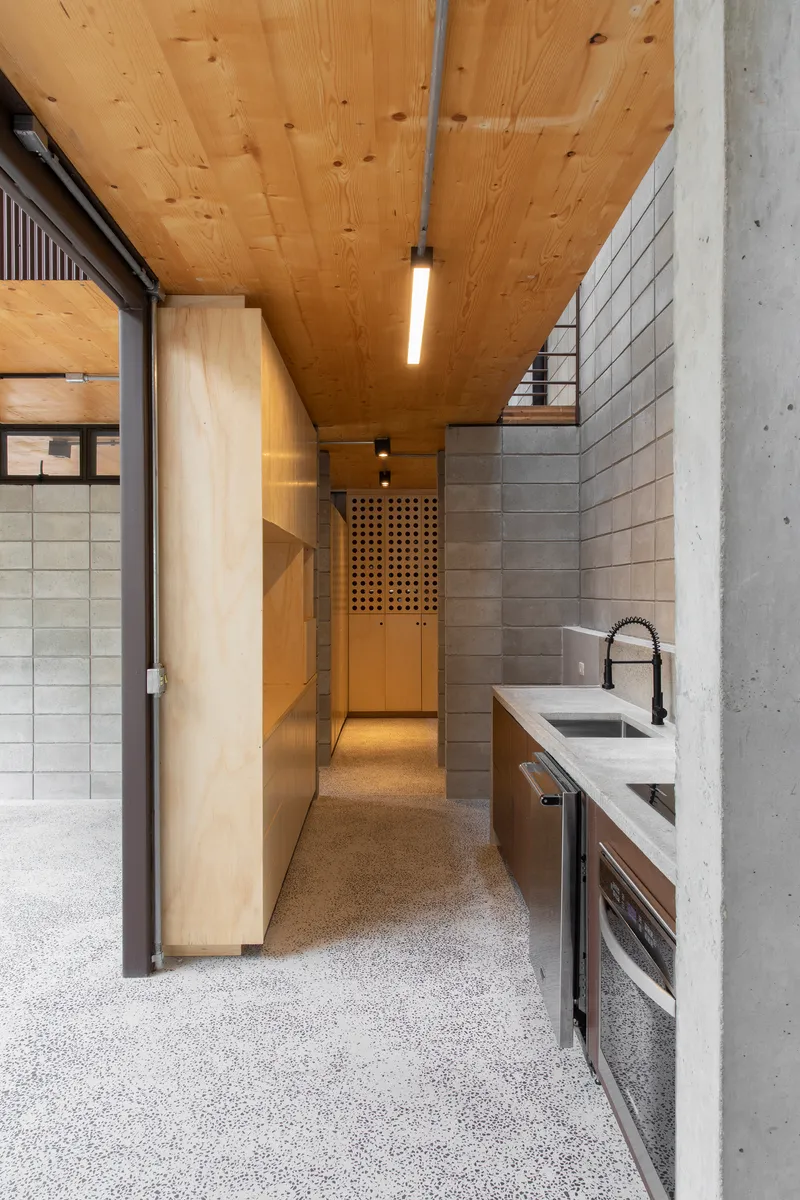 Galley kitchen with terrazzo flooring, plywood wardrobes and concrete block walls beneath a timber plank ceiling