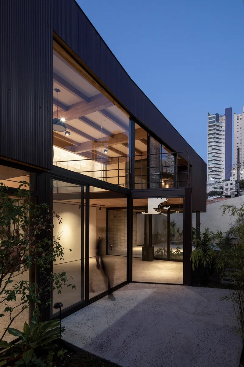 Glazed facade with black steel frames and corrugated metal cladding illuminated at dusk with tower beyond