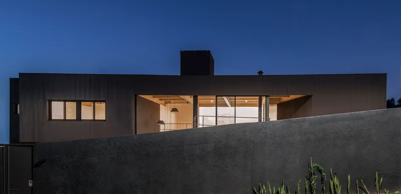 Dark corrugated metal facade with illuminated interior visible through large glazed openings at dusk