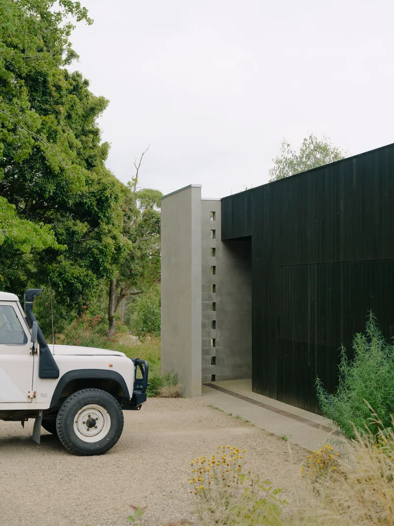 Concrete wall with vertical window openings meeting corrugated metal cladding beside a parked vehicle