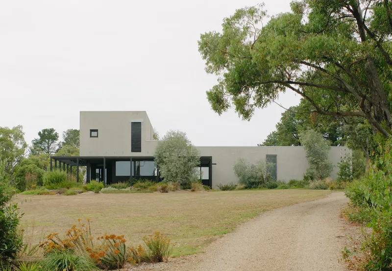 Concrete facade with raised volume and glazed lower level framed by native trees and gravel driveway