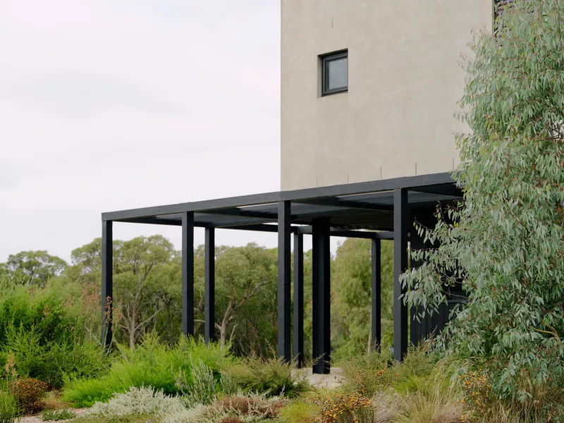 Black steel pergola extending from the concrete tower wall into planted garden beds
