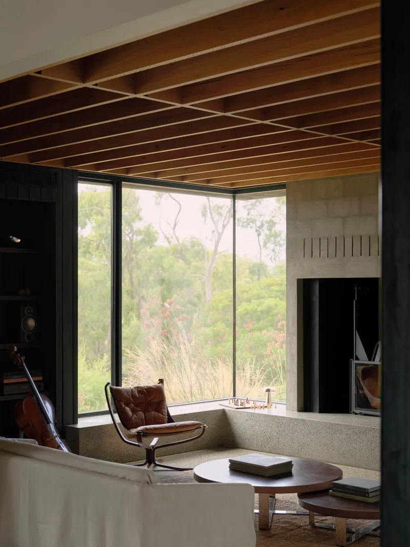 Living room with timber slat ceiling and floor-to-ceiling windows overlooking bushland with cello beside fireplace