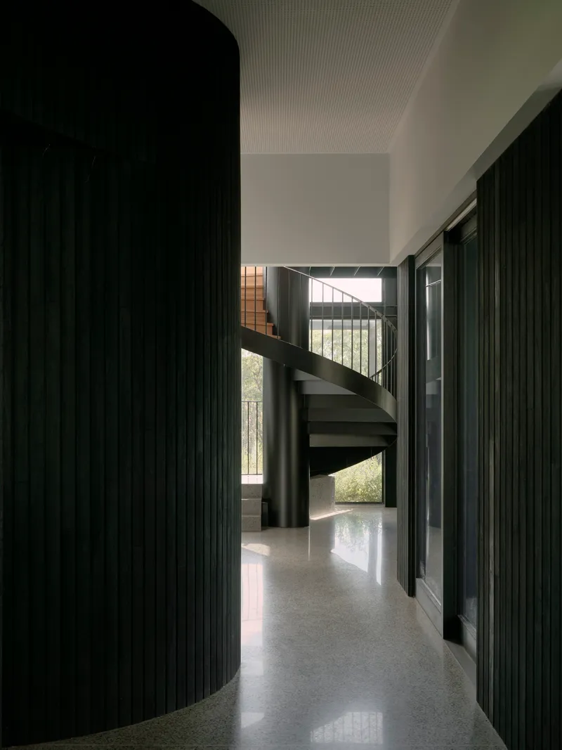 Curved black timber-clad wall beside a spiral staircase in a corridor with polished concrete floors