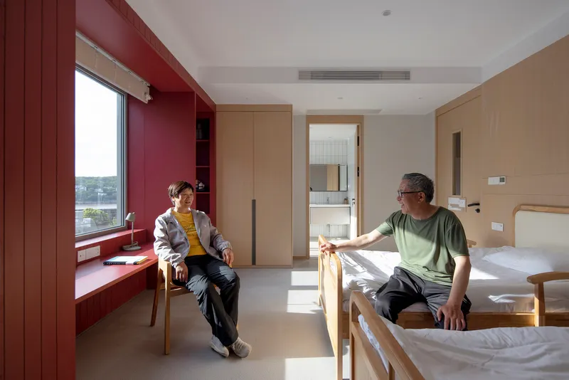 Shared room with two timber beds and a maroon window niche where two people sit in afternoon light