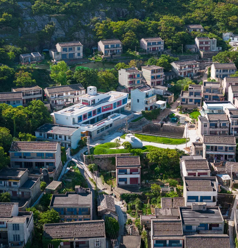Aerial view of white volumes with colored window bands nestled among traditional tiled-roof houses on a hillside