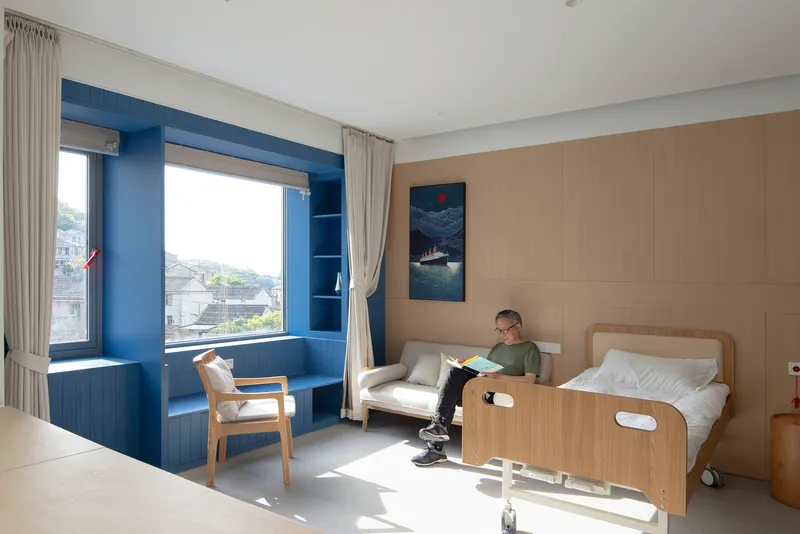 Patient room with timber wall panels, blue window alcove, and a person reading beside the bed