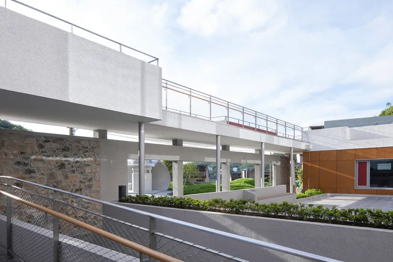 Elevated concrete walkway on cylindrical columns spanning over a stone retaining wall and landscaped courtyard