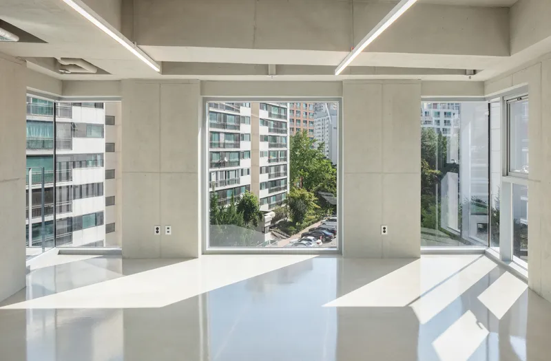 Interior space with exposed concrete ceiling beams and floor-to-ceiling windows overlooking neighboring buildings