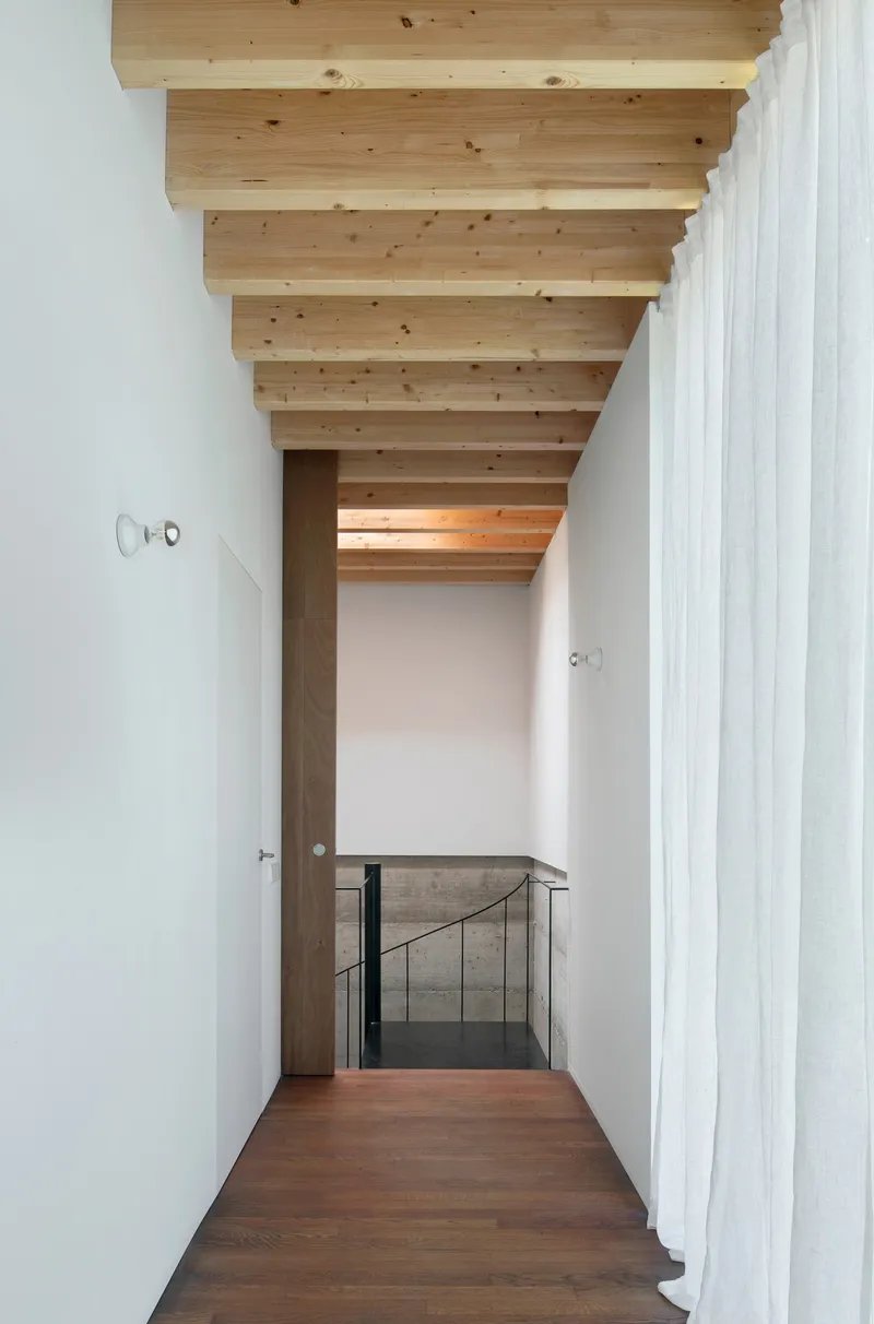 Corridor with exposed timber ceiling beams leading to glass-railed stairwell with white walls and timber flooring