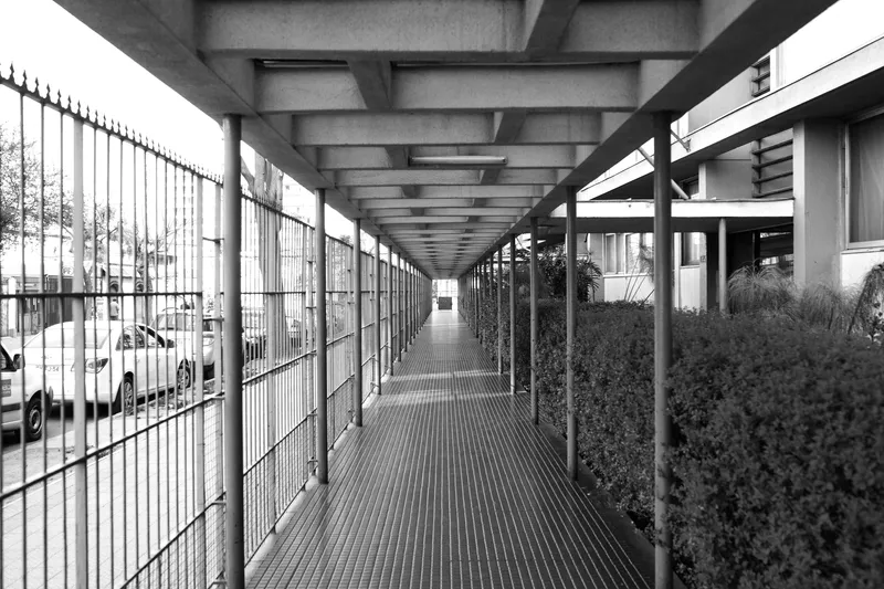 Long covered walkway with coffered concrete ceiling, metal fence and hedge along parked cars
