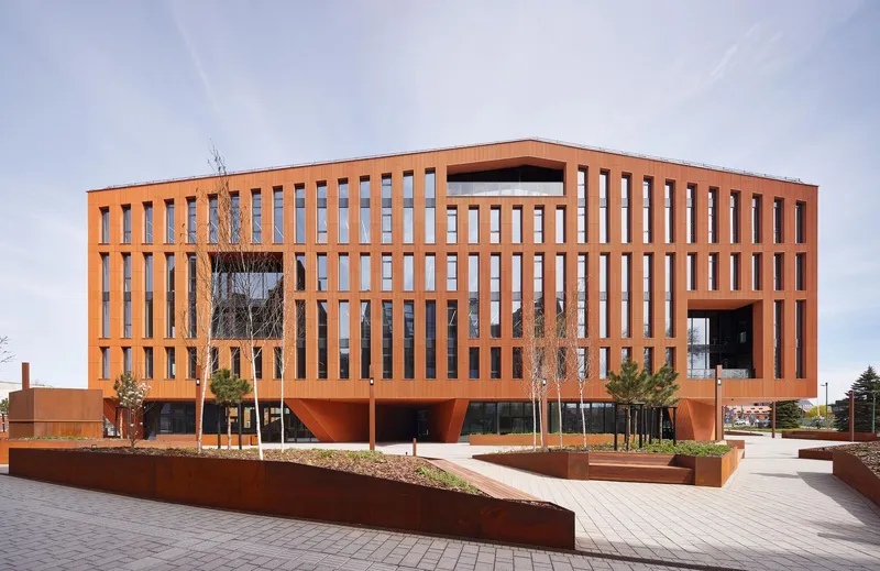 Plaza view of the terracotta facade with vertical slot windows and elevated volume above planted beds