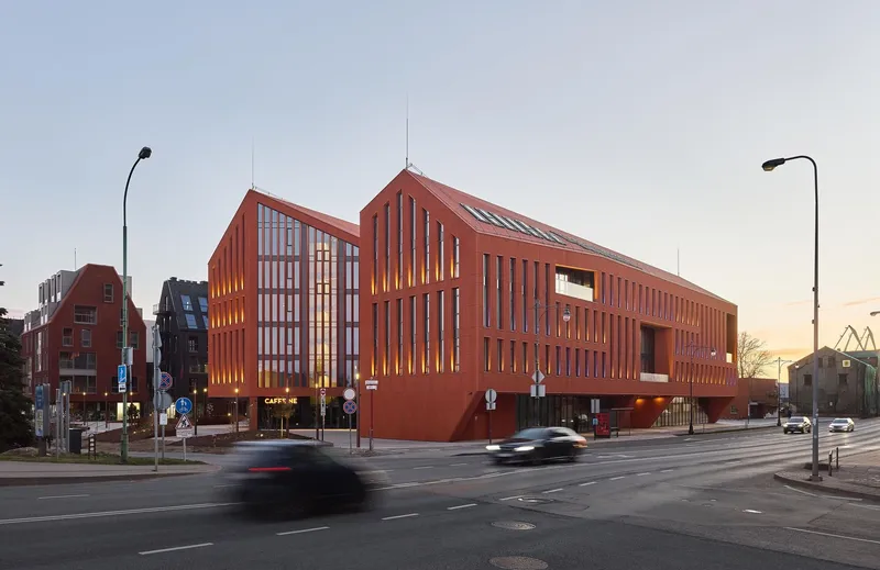 Street corner view of the red brick facade with vertical fenestration at dusk with passing cars