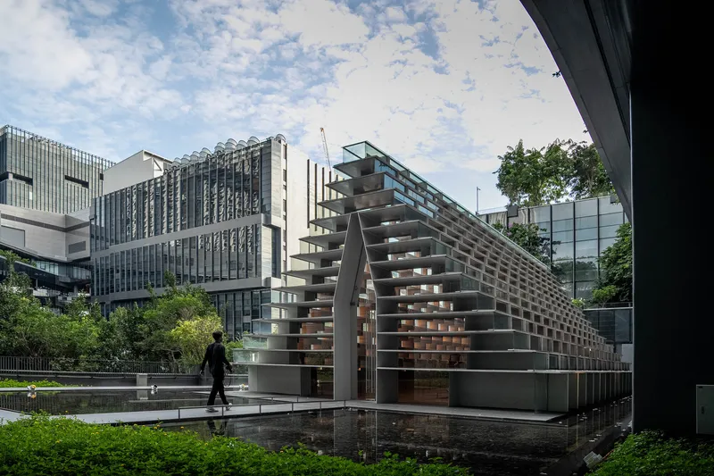 Pyramid-shaped pavilion with open slat facade beside a reflecting pool as a person walks past