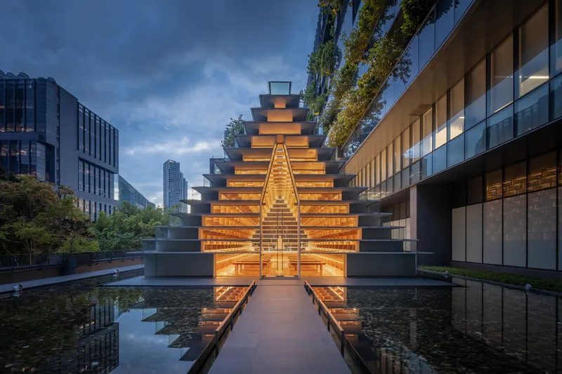 Stepped pyramid pavilion illuminated at dusk, flanked by reflecting pools and surrounded by glass office buildings