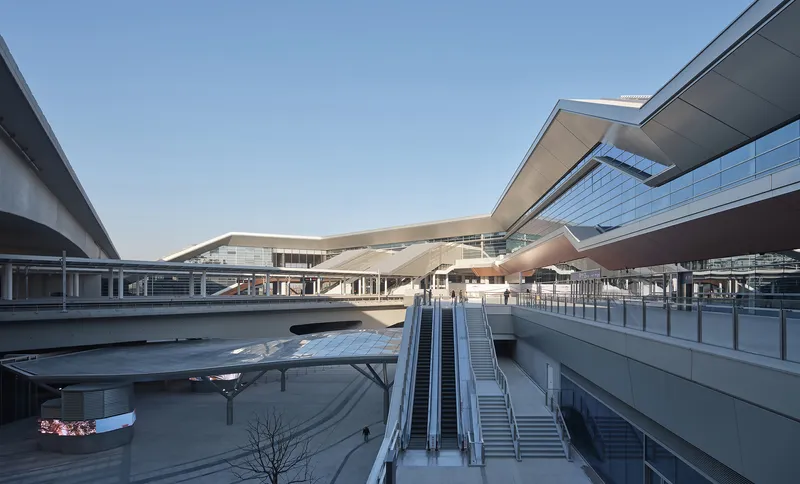 Multilevel transit plaza with escalators leading between concrete platforms under a clear sky