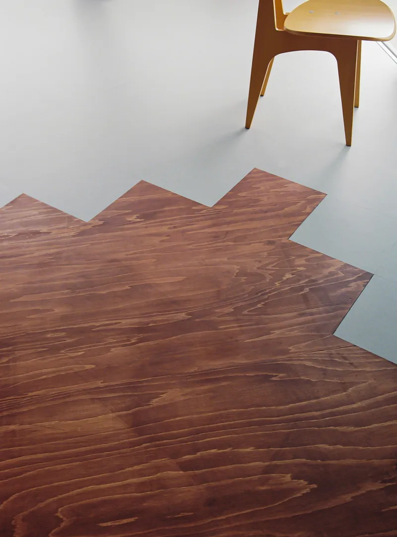 Overhead view of zigzag wood floor edge meeting pale linoleum with timber chair legs visible