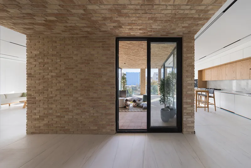 Interior view through black-framed glass doors to an exterior terrace with woven brick ceiling and potted plants