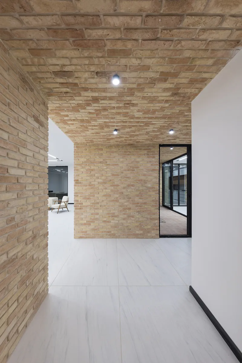 Corridor with textured brick wall and ceiling meeting white plaster surfaces under recessed lighting