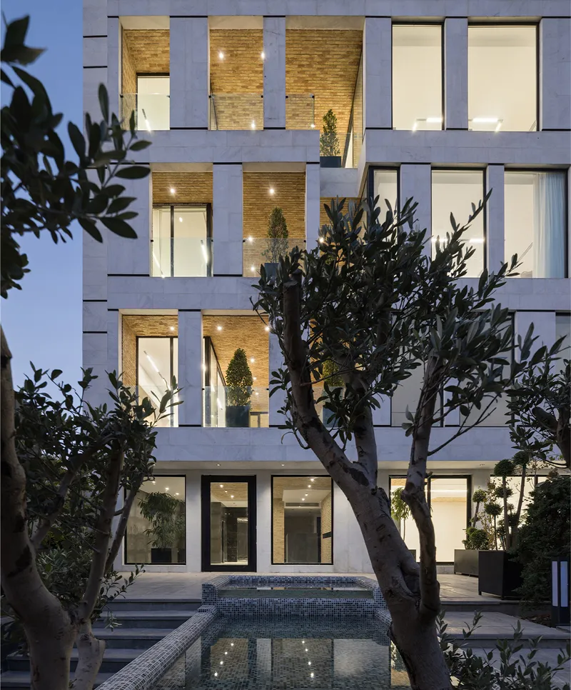 Street facade at dusk showing staggered brick balconies framed by concrete slabs behind mature olive trees