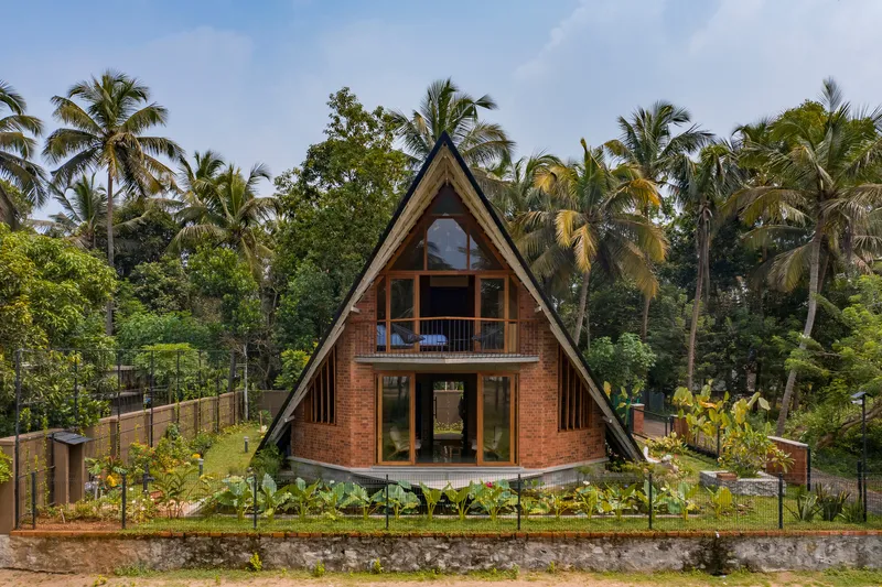 Front elevation showing the triangular gabled facade with exposed brick walls and upper balcony framed by coconut palms