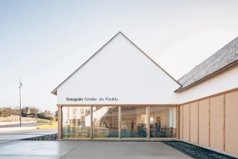White gabled volume with full-height glazing revealing the interior workshop space beneath an overcast sky