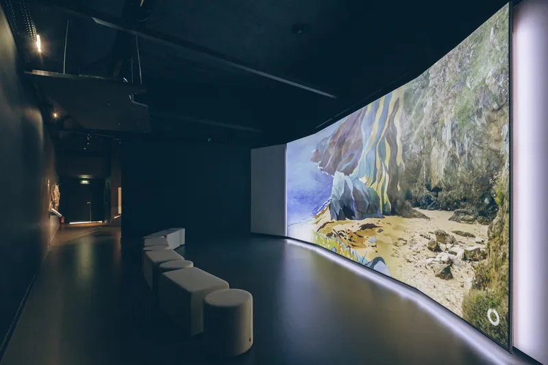 Darkened screening room with curved projection wall displaying coastal imagery and a row of white benches