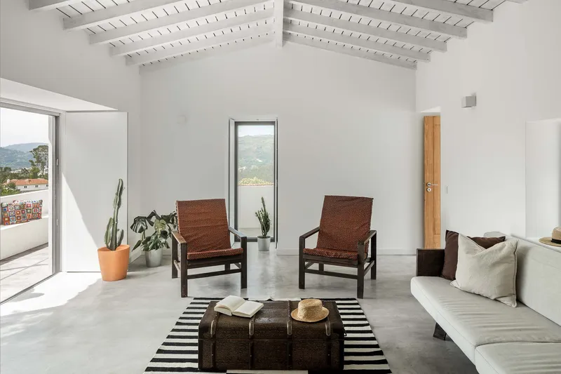 White living room with exposed timber ceiling joists and two woven chairs facing tall vertical windows