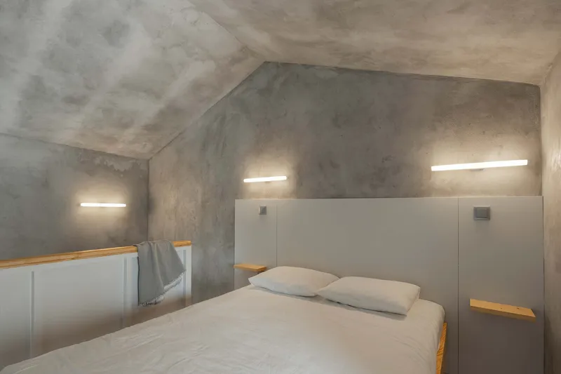 Bedroom with sloped concrete ceiling, horizontal recessed lighting, and grey paneled headboard with timber shelving