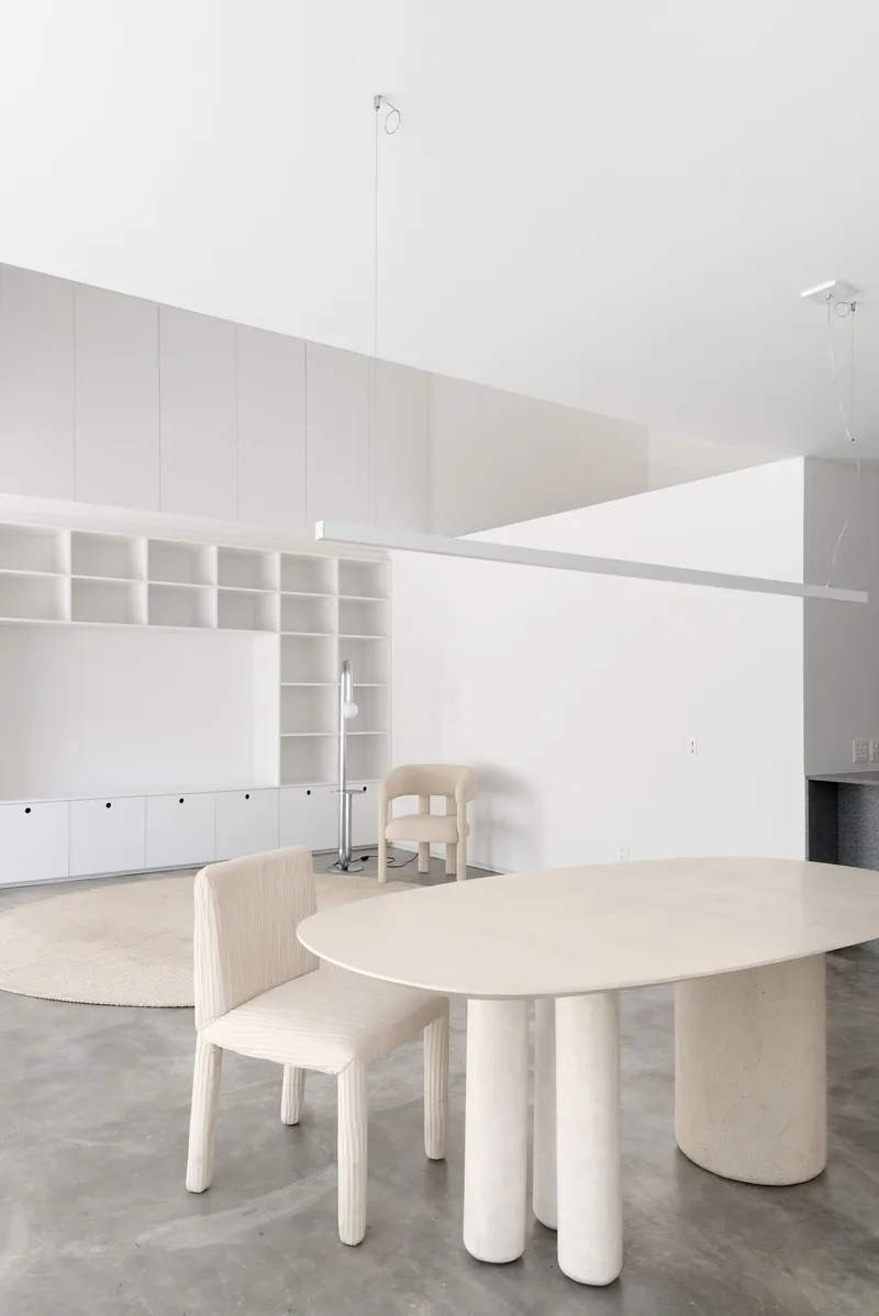 Light wood dining table with cylindrical concrete legs surrounded by wooden chairs under vaulted white ceilings