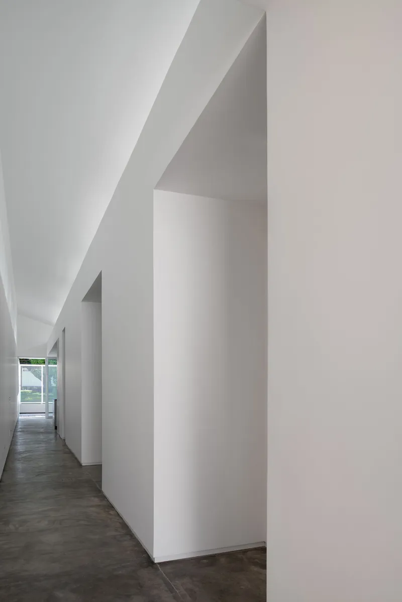 Narrow corridor with angular white walls and sloped ceiling leading toward a distant window