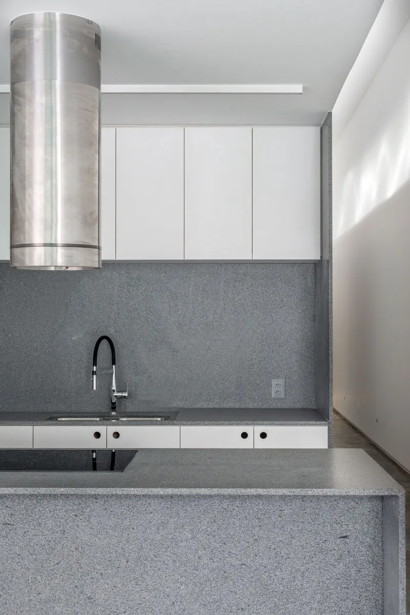 Kitchen with grey terrazzo countertops, white cabinets, cylindrical steel range hood, and sunlight entering from the right