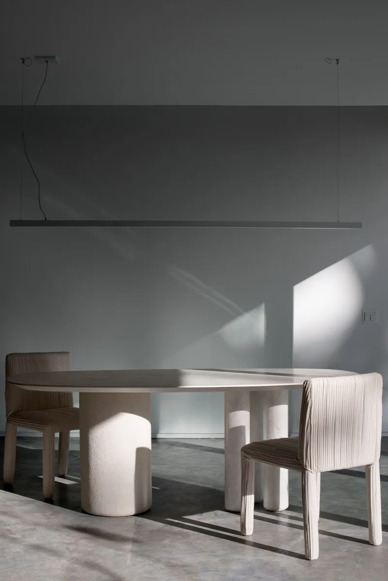 Dining area with circular table, ribbed chairs, and overhead linear pendant casting shadows on the wall
