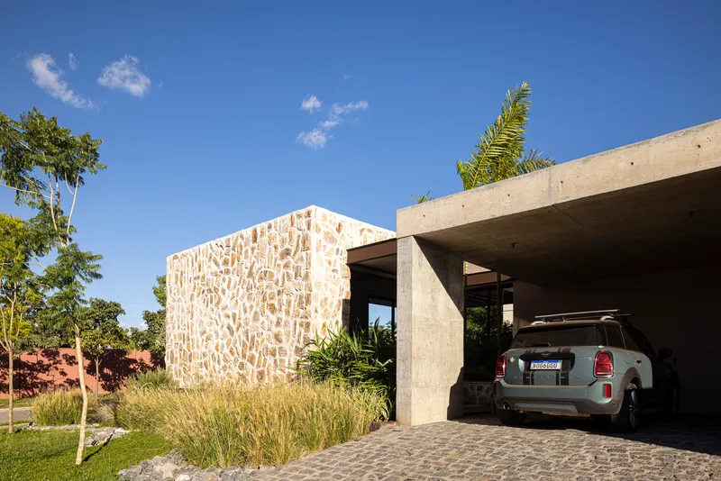 Textured stone facade volume adjoining a concrete carport canopy under afternoon sunlight