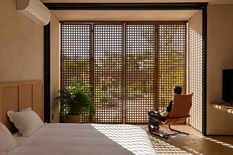 Bedroom with perforated wooden sliding doors filtering dappled sunlight onto a person seated in a lounge chair