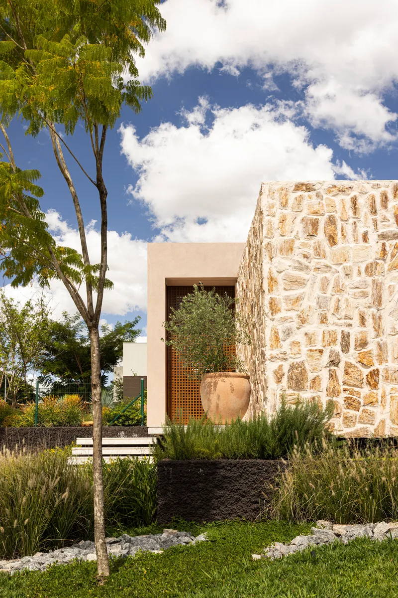 Entry facade with rough limestone wall, perforated timber screen, and planted garden beds
