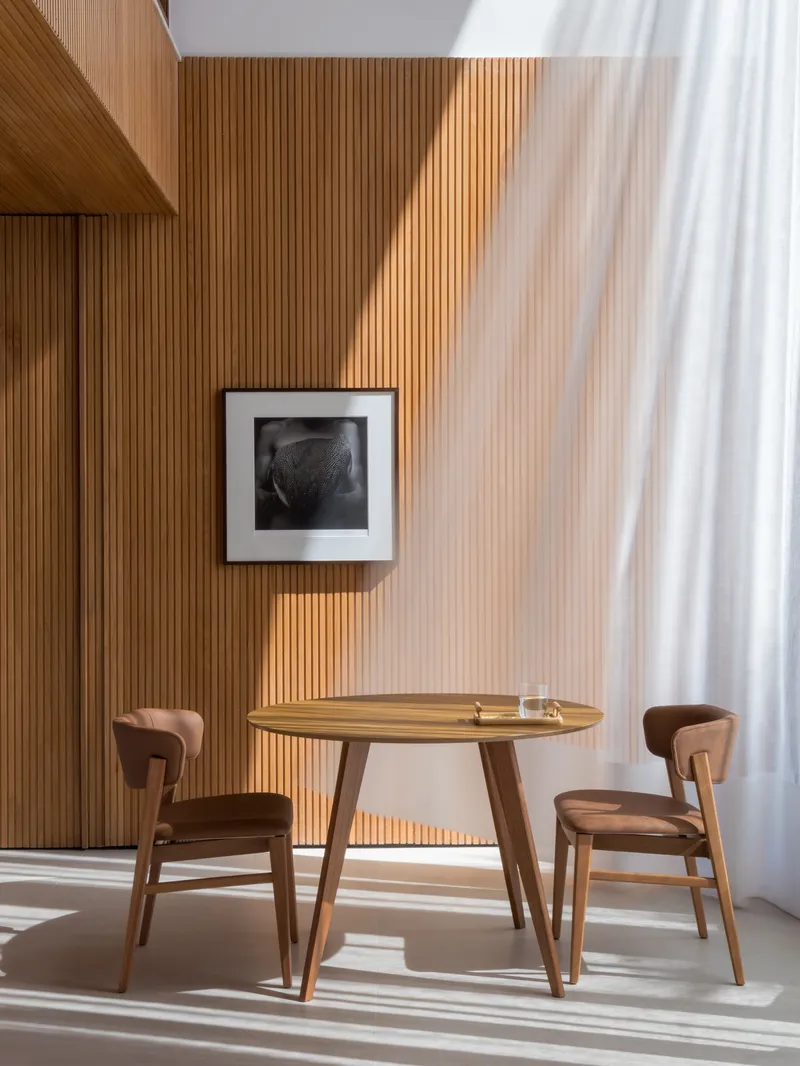 Dining area with round timber table and chairs against slatted wood wall in morning light