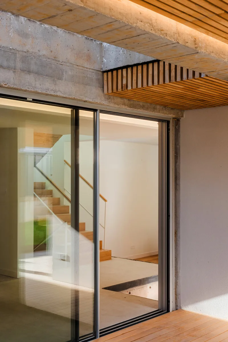 Exposed concrete beam with timber slat ceiling and open sliding glass door revealing internal staircase