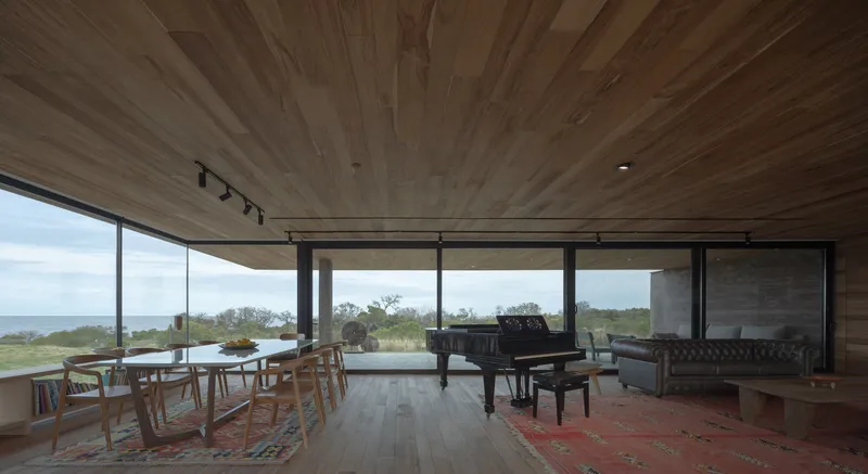 Interior living space with timber-clad ceiling, grand piano, and floor-to-ceiling windows overlooking coastal terrain