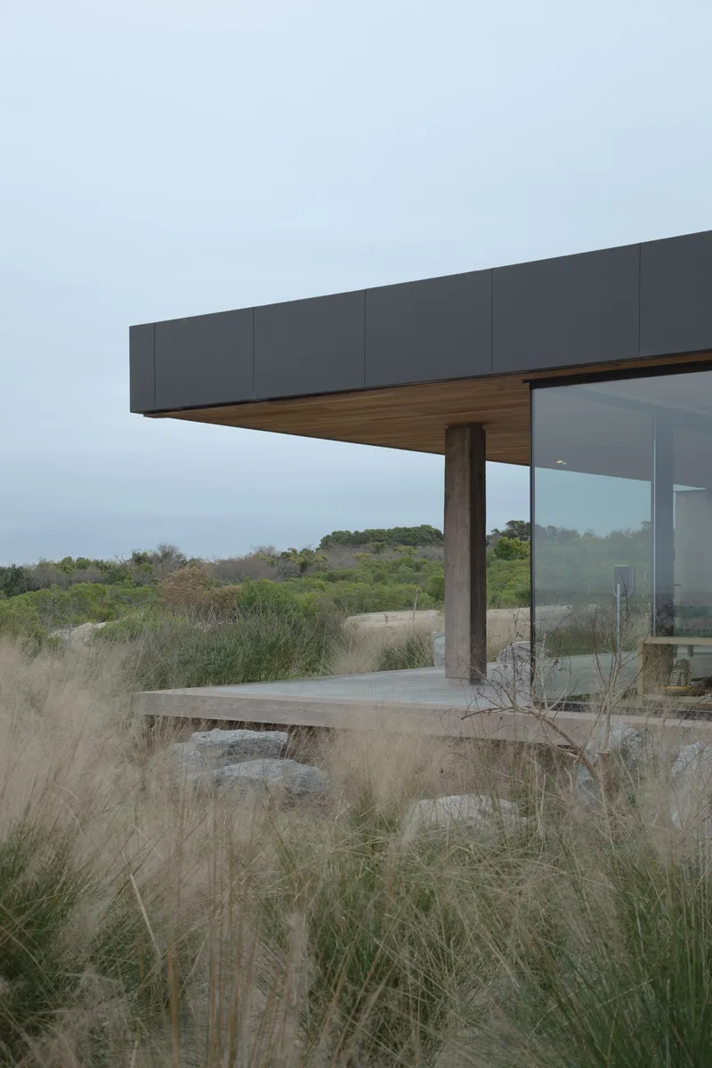 Cantilevered roof edge with timber column and concrete bench surrounded by dry coastal grasses under overcast sky