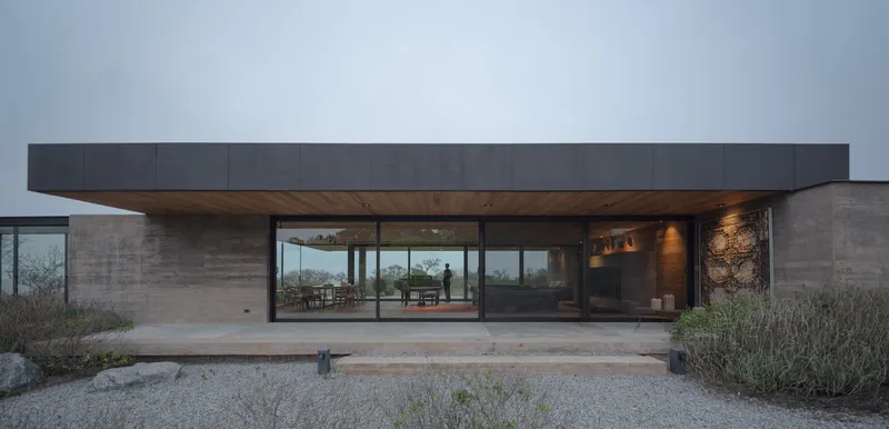Front facade with recessed glazing beneath a cantilevered dark metal roof overhang and rammed earth walls