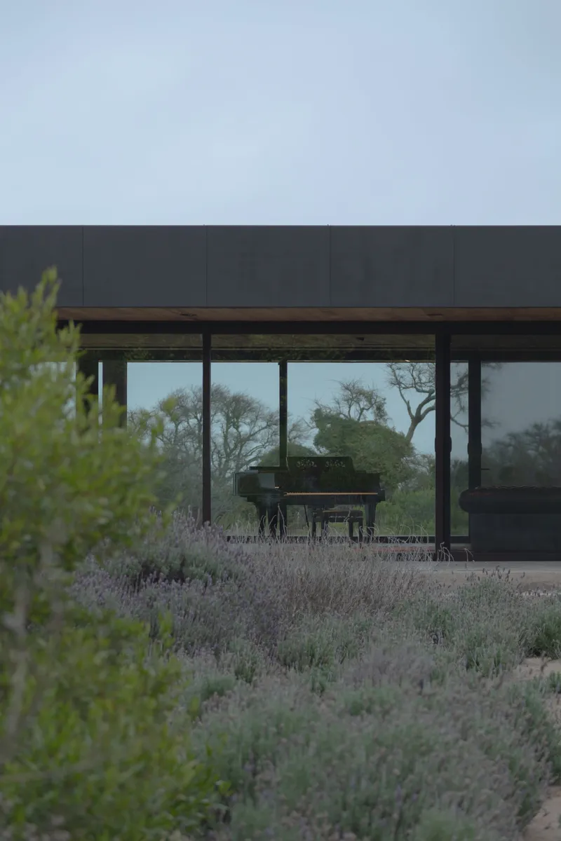 View through floor-to-ceiling glass walls to a grand piano surrounded by low desert grasses