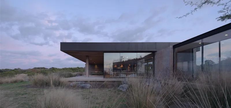 Cantilevered roof sheltering glass-walled living spaces set among grassland at twilight