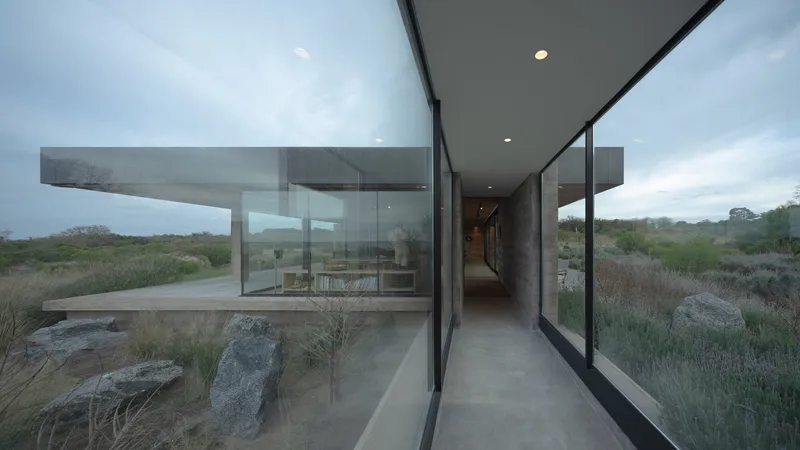 Glass-enclosed corridor with exposed rammed earth wall and overcast sky beyond the landscape