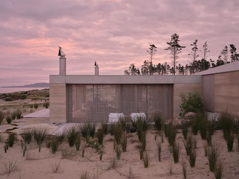 Coastal pavilion with vertical timber cladding and white chimneys surrounded by beach grasses at dusk