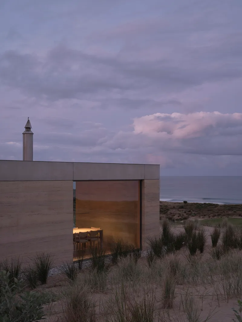 Glass corner room with illuminated interior and coastal view beyond dune grasses at twilight