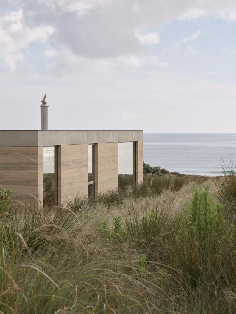 Concrete facade with vertical timber screen openings seen through coastal dune grasses overlooking the ocean