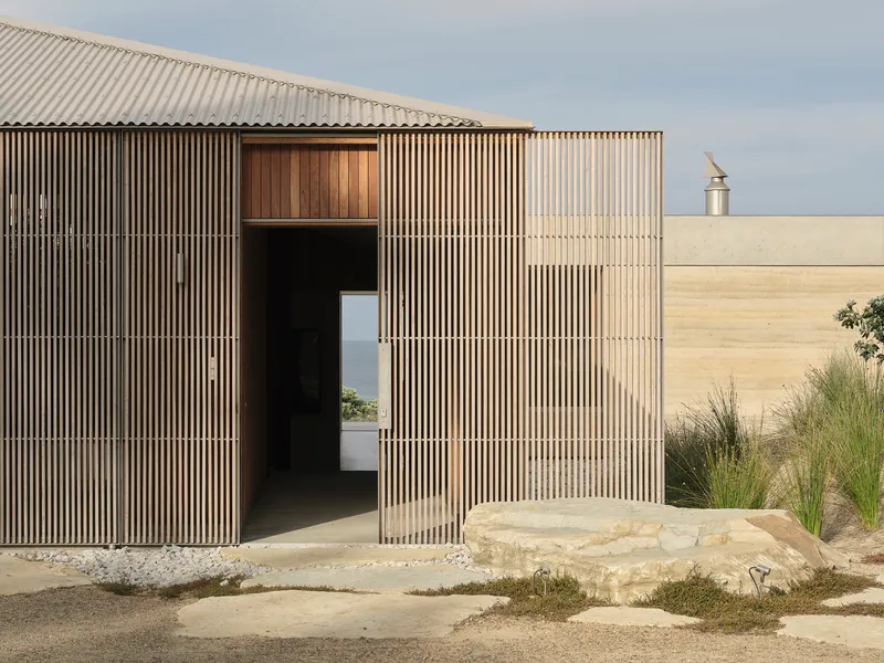Entry pavilion clad in vertical timber battens with pivoting door and limestone outcrop beside gravel courtyard