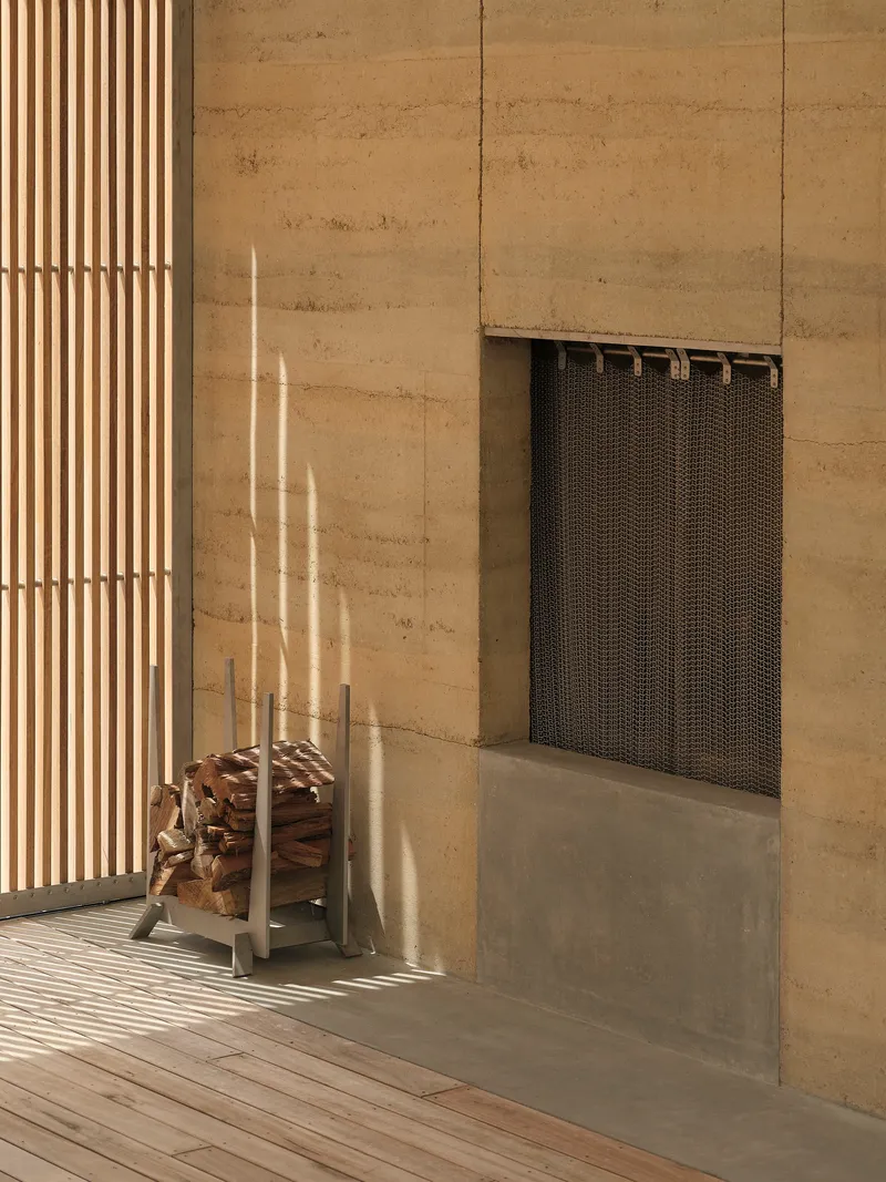 Fireplace recess with metal mesh screen in rammed earth wall beside timber slatted screen in afternoon light