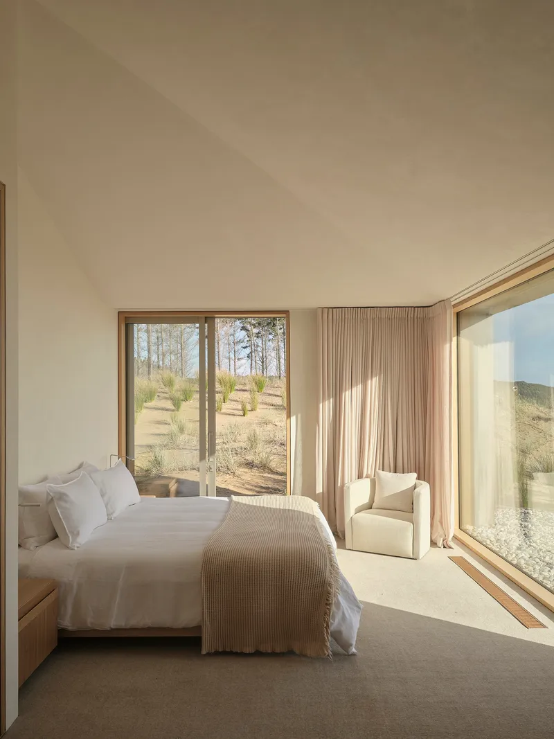 Bedroom with floor-to-ceiling glazing overlooking grassy landscape with scattered trees under soft daylight
